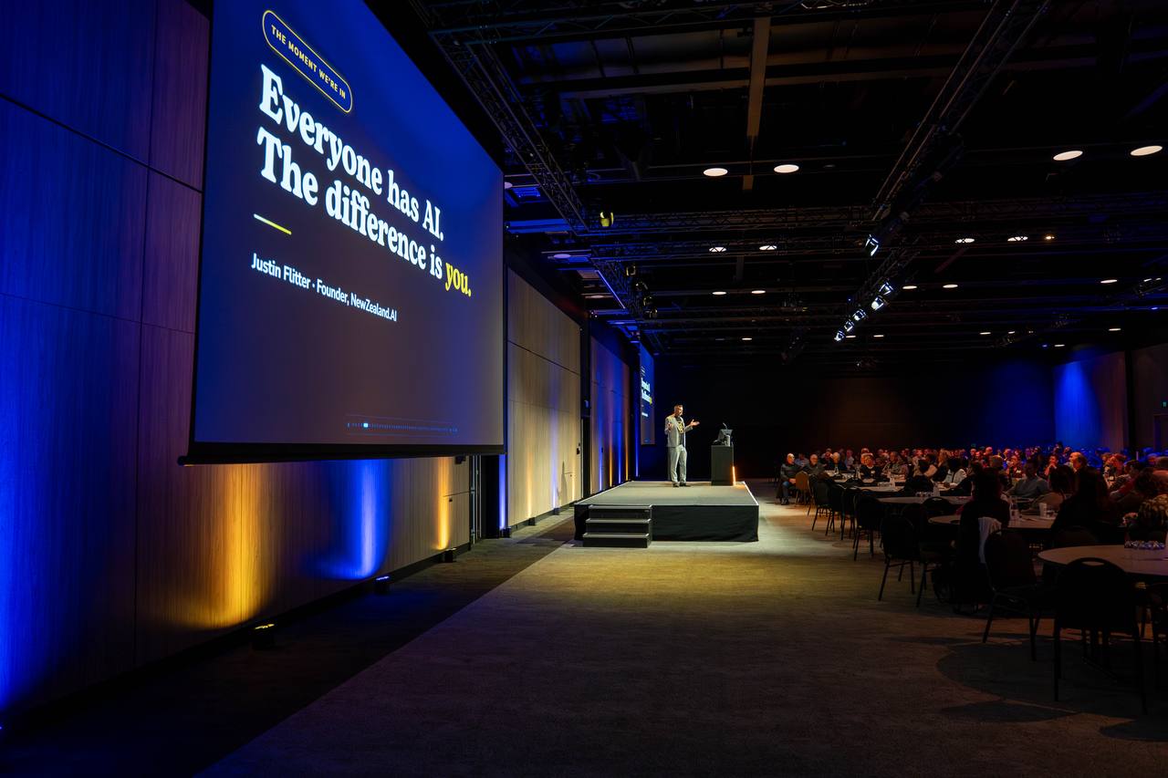 Justin Flitter speaking on stage to a seated audience, beside a screen reading “Everyone has AI. The difference is you.”